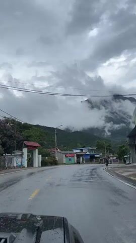The camera is mounted on a vehicle driving on a wet, paved road through a small village nestled near a mountain range. The mountain peaks are partially obscured by low-hanging clouds. Buildings, including simple houses and a business, line the road. Electrical poles run along the roadside. A motorcyclist can be seen traveling in the same direction. The sky is overcast.