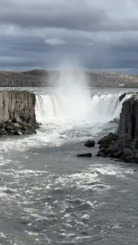 The video captures a wide shot of a powerful waterfall under a cloudy sky. A large volume of grey water cascades over a wide, dark rocky cliff, creating a significant amount of white mist that rises above the falls. The river below the waterfall is turbulent and frothy, filled with rapids as the water flows away from the main drop. Dark, jagged rocks and cliffs line both sides of the river and the waterfall. In the distance, beyond the falls, more rugged, dark land formations are visible under the overcast sky. No people or text are visible in the scene. The overall impression is one of raw natural power and a dramatic, rugged landscape, typical of the Icelandic environment mentioned in the context.