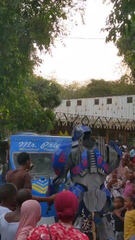 The video shows a person in an Optimus Prime costume posing with a crowd of children. The person, who is standing on their own, is wearing a full-body suit, the primary colors of which are blue and gray. The crowd is a mix of genders and ages. Most people appear to be of African descent, based on skin tone. The crowd is gathered around the Optimus Prime figure, with some children reaching out to touch the costume and others taking photos with their phones. The crowd is standing in what appears to be an outdoor recreational area with trees on either side. Behind the person in the costume and the crowd is a bright blue food truck with the words "Mr. Price Bullet Train" printed on it. In the background, there is a white, boxy structure that resembles an old shipping container that has been turned into a building. A wrought iron fence or awning spans its front. The sky is bright, suggesting it is daytime. The overall scene is lighthearted and positive, with the crowd enjoying the presence of the Optimus Prime character.