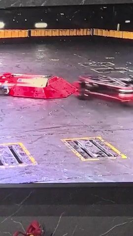 The video shows a robot combat competition. Two red robots are actively engaged in a fight on a circular arena floor. One robot, appearing damaged and red, is on its side. The other robot, also red and in motion, is facing the damaged robot. The arena floor is gray with yellow markings and what appear to be grates or hazards. In the background, spectators are visible in the stands. Intercut with the fight footage are shots of a person wearing a black baseball cap, glasses, and a gray and black shirt, holding a remote control, and standing next to a transparent enclosure containing robot parts. The person appears to be a robot operator or team member. The overall scene depicts an intense robot battle.