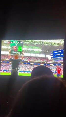 A soccer match is being shown on a television. The score is tied 1-1. A player in a red jersey with blue and white accents, number 5, is on the field. Another player in a red jersey with blue and white accents approaches him and puts an arm around his shoulder. In the background, a stadium filled with spectators is visible. A person's head, wearing a dark hat, is in the foreground, obscuring the bottom of the screen. A man is holding up a digital board that displays "88:98" and "Ref".