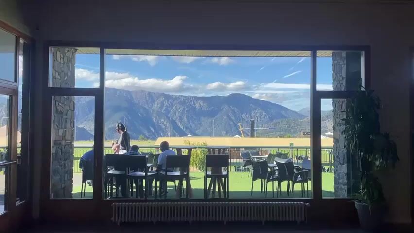 The video shows a view from inside a building looking out through a large window onto a patio area and beyond to a mountain range. The interior has stone columns on either side of the window and a plant on the right. Inside, there are several tables and chairs. Three people are seated at one table, and one person is standing and appears to be interacting with them. On the patio, there are more tables and chairs set up on a grassy area. In the background, there is a flat area with some structures, possibly an airport or sports field, and then the large, rugged mountains under a bright blue sky with scattered white clouds. The sun is casting shadows, indicating it is daytime. The overall impression is a scenic view from a restaurant or lounge.