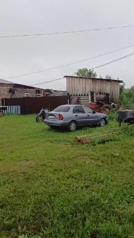 The video shows a view of a rural residential area on a cloudy day. In the foreground, there is a grassy yard with some debris. A gray sedan and a motorcycle are parked in the yard, with a small wooden structure in the background. Further back, a house with a brick facade and a gray roof is visible, along with a turquoise fence and a pile of dirt. A power pole and wires are also present. The camera pans to the right, showing a gravel driveway leading to a garage or outbuilding. Another house with a red car parked in front of it is then seen, with a red fence surrounding the property.