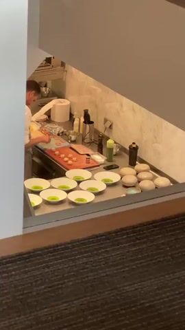 This media file provides a glimpse into a professional kitchen environment, likely at a restaurant in The Hague, Netherlands. The scene is set on a stainless steel countertop, illuminated by overhead lighting, suggesting it is either daytime or evening within a well-lit establishment.

The primary subject is a chef, seen from the side, wearing a white chef's coat and a grey apron. He is actively engaged in preparing food on a red cutting board. Several small, circular food items, possibly appetizers or components of a dish, are arranged on the board. Next to them are various kitchen tools and ingredients, including bottles of sauces, a blender, and what appears to be some sort of meat or fish. A large roll of paper towels is also visible.

In the foreground, a collection of white, shallow bowls, each containing a vibrant green liquid, is neatly arranged. These are likely prepped for serving. To the right of the bowls, a group of smooth, pale, oval-shaped objects are stacked, their purpose unclear but possibly decorative or part of the plating. A few other smaller dishes and a smartphone are also present on the counter.

The overall atmosphere in the kitchen appears to be one of focused preparation. The chef's posture suggests concentration on his task. The clean and organized state of the kitchen implies a high standard of operation.

The media file also offers a wider view of the restaurant's interior. Through a glass partition, patrons can be seen seated at tables in a dining area, suggesting the kitchen is part of a larger, operational restaurant. The interior design appears modern and sophisticated, with elements of light wood flooring and clean white walls. The time of day is not explicitly evident from the weather or natural light, but the general ambiance suggests a service period. No visible text is present in the media.