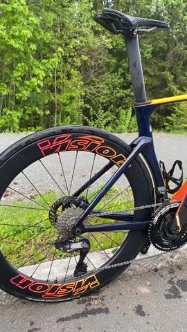 The video shows a Cannondale road bike parked on a street. The bike has a navy blue, pink, and orange frame with Vision wheels. It's resting against a curb next to a road with greenery in the background. The sky is overcast. The camera pans around the bike, showcasing its features.