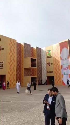 The video captures the exterior of a large exhibition hall, likely Al Machouar – Stinia, Morocco, during the International Agricultural Exhibition of Morocco (SIAM).

The prominent entrance features a modern building with facades made of textured panels resembling wood, decorated with stylized wheat stalk patterns in various shades of brown, orange, and beige. Some panels integrate large LED screens displaying graphics, while others have open display boxes, one of which appears to house a small animal sculpture.

Large signage on the building clearly states "SIAM 18" (referring to the 18th edition of the event) and "SALON INTERNATIONAL DE L'AGRICULTURE DU MAROC" in both French and Arabic, along with the dates "20 AU 28 AVRIL 2024". It also notes the event is "Under the High Patronage of His Majesty King Mohammed VI." Moroccan and Portuguese flags are displayed near the entrance, indicating participating nations.

Throughout the video, numerous people are seen walking towards and away from the entrance. Attendees are diverse, including men and women in both modern casual and business attire, as well as some individuals wearing traditional North African clothing like djellabas. Many appear to be purposeful, some looking at their phones.

The scene depicts a bright, overcast daytime, with diffused light. The ground is paved with cobblestones. In the broader shots, palm trees and several Moroccan flags are visible in the distance, set against a backdrop of a city skyline that includes buildings and a prominent tower or minaret, reinforcing the Moroccan setting. The overall atmosphere is one of moderate activity at a public event.
