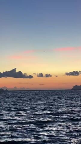 The video shows a tranquil ocean at sunset. The sky is a gradient of blue, orange, and pink, with a few scattered clouds. The ocean is dark blue and slightly rippled. In the distance, silhouettes of islands can be seen against the horizon. The overall atmosphere is peaceful and serene.