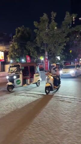 A nighttime scene on a paved road shows several vehicles. On the left, a white tuk-tuk with a green Grab logo is driving towards the viewer. Its headlights are on. Further back and to the right, a scooter with two people on it is also moving towards the viewer. The person in front is wearing a helmet and the person behind them is also wearing a helmet. To the right of the scooter, a white car with its headlights on is visible. In the background, trees with lights and buildings are present. The road surface is wet, reflecting the vehicle lights. Another tuk-tuk is parked on the side of the road behind the moving tuk-tuk.
