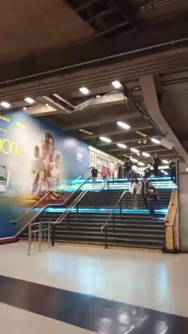The video captures a scene inside an underground transit station, likely in Providencia, Chile. Numerous people are seen ascending and descending a wide set of stairs equipped with modern, color-changing LED lighting along the steps, transitioning from yellow to blue. To the left of the stairs, a large advertising poster for the "HBO ORIGINAL" series "Euphoria" is prominently displayed, also featuring the "HBO Max" logo and the word "PIEDAD." In the background, a train appears to be either arriving at or departing from the platform. The station features a functional, industrial ceiling with visible pipes and fluorescent lighting. Metal railings line the stairs, and the floor is a light-colored tile with darker accent strips. The overall impression is a busy, well-lit commuter environment.