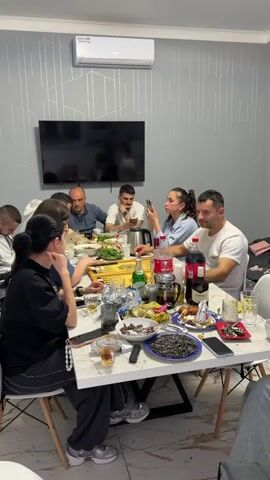 A group of ten people, a mix of men and women, are sitting around a white marble table. The table is cluttered with food, drinks, glasses, plates of sunflower seeds, and personal items like phones and wallets. In the foreground, two women are playing backgammon on a wooden board. The women are focused on the game, with one of them smiling. The other people at the table are either watching the game, looking at their phones, or engaged in conversation. 

The room has light grey walls decorated with geometric patterns. Above the table, there's a wall-mounted air conditioner and a television. A white chair is partially visible on the right side of the frame. The lighting in the room appears to be bright and even.