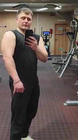 The video shows a man standing in a gym, taking a selfie. He is wearing a black sleeveless shirt, black sweatpants, and light-colored sneakers. The man flexes his left arm. He has short, light-colored hair. The gym has various exercise machines visible in the background, including a pull-up bar, an elliptical machine, and a weight lifting machine. The floor is red with square tiles. A door with a green sign above it is visible in the background.