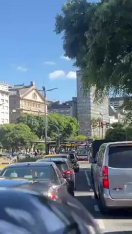 The video shows a view from inside a moving car in Buenos Aires, Argentina. First, a tall building with "NH HOTELS" on top and a green bus are visible. Then, a group of people are seen walking on a patch of green space, partially obscured by foliage. Next, another building with a dome and glass roof is visible. Finally, the Obelisco de Buenos Aires comes into view, framed by buildings and billboards under a clear blue sky. Traffic and city sounds are audible.