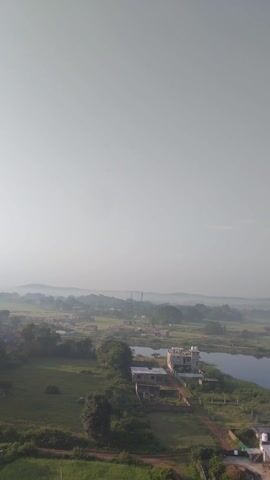 This is a drone's-eye view, captured vertically, of a rural landscape. The scene is dominated by a vast expanse of pale blue sky taking up the left side of the frame. The right side reveals a body of water, likely a river or large stream, snaking through a lush green, somewhat overgrown landscape. The banks of the water are lined with vegetation. In the middle ground, there are several small buildings, some appearing under construction or in a state of disrepair, with visible concrete structures. Further in the distance, the landscape appears to be a patchwork of fields and scattered trees, with a hazy, muted green and brown color palette. A few structures, possibly houses, are visible in the far distance. The overall impression is of a rural or semi-rural area, possibly with some industrial or agricultural activity. There are no people visible in the footage.