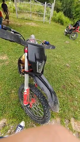 A person's hand, wearing a blue shirt and white sneakers, is visible in the foreground of the video, obscuring the bottom half of the frame. The hand is holding onto a black, red, and silver dirt bike, which is positioned in the center of the frame. The dirt bike has knobby tires, a black headlight, a yellow gas tank, and red accents on the suspension and wheels. The bike is parked on a grassy area. In the background, a white metal fence is visible, and beyond that, trees and foliage create a natural backdrop. There are two other dirt bikes in the background. One is red and parked to the right, and another is orange and appears to be in motion further to the right. Two people are partially visible in the background, standing near the fence on the left side of the frame. One is wearing a black shirt and appears to be holding a shovel.