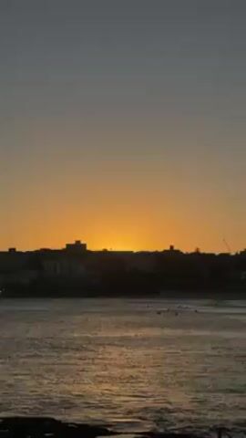 The video shows an ocean view in a coastal city at dusk, with the sun setting in the background. The sky is a gradient of orange and gray, with the sun casting a warm glow on the water. There are several silhouetted buildings on the coastline. In the water, there are people swimming or surfing. The water is dark and reflective. The foreground has dark rocks.
