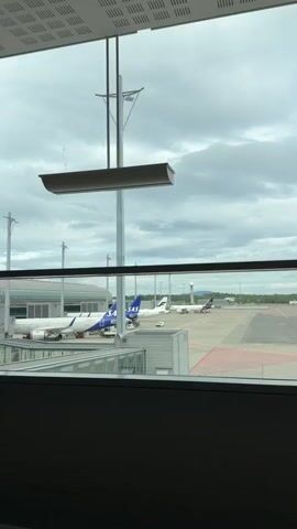 The video is filmed from inside an airport terminal, looking out through a large window at an airport tarmac. Several airplanes are visible, parked at gates and on the tarmac. One prominent airplane in the foreground has the SAS logo and blue and white livery. Another airplane with white and blue livery is parked next to it. The sky is overcast with clouds. A jet bridge connects to one of the airplanes. The interior of the airport terminal is visible in the foreground, including a section of a ceiling with ventilation grates and a dark, opaque surface at the bottom of the frame. A long, rectangular light fixture hangs from the ceiling by wires, obscuring part of the view of a tall pole with lights and wires on the tarmac. The camera appears to be stationary or panning very slowly.