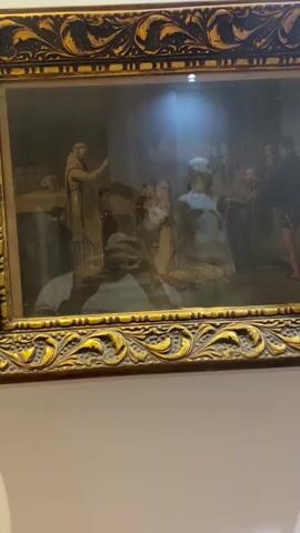The video shows a framed artwork hanging on a wall. The artwork appears to be a painting or a diorama with figures. In the foreground, there is a reflection of a person holding a phone, taking the video. The artwork itself depicts a scene with several figures, including one in a monk-like robe addressing a group of people. There are also statues or figures of a nude man and a woman. The environment appears to be indoors, possibly a museum or gallery, with a spotlight illuminating the artwork from above. The frame of the artwork is ornate and golden. The reflection of the person taking the video obscures parts of the artwork.
