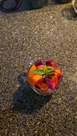 The video shows a close-up of a clear glass cup filled with diced fresh fruit, placed on a speckled granite countertop. The fruit mixture prominently features orange cubes, likely papaya, and vibrant pink pieces with black seeds, characteristic of dragon fruit. A garnish of fresh green herbs, possibly basil or mint, rests on top of the fruit.

Initially, the fruit cup is stationary on the countertop. A thin, light-colored stream of liquid begins to pour into the cup from above. As the video progresses, the pouring action becomes clearer, revealing a white, creamy liquid, consistent with condensed milk, being dispensed from a silver-colored can. The stream of liquid is directed over the fruit and herbs, coating them. The pouring continues until a noticeable amount of the creamy liquid covers the fruit, enhancing its appearance.

The scene appears to be an indoor setting, likely a kitchen, given the countertop and the activity. The lighting is moderate, suggesting ambient indoor illumination. No individuals are visible in the video. The preparation of fruit with condensed milk is a common culinary practice in places like Vietnam. There is no visible text on any objects in the frame.