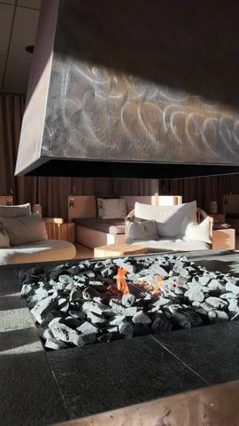This video shows a close-up shot of a modern indoor fireplace with a large metal hood. The fireplace is filled with dark stones and has a small fire burning in the center. In the background, a lounge area is visible with comfortable seating, including sofas and cushions. The room is dimly lit, with warm ambient lighting and shadows cast by the sunlight streaming in from the side. The overall atmosphere is cozy and inviting.
