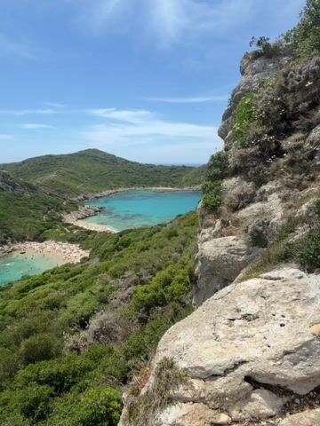 The video shows a panoramic view of a horseshoe-shaped bay with clear turquoise water and a sandy beach. Lush green vegetation covers the surrounding hills, which are rocky and dotted with small white flowers in the foreground. A few scattered clouds drift across the bright blue sky. The beach is populated with numerous people, some swimming or sunbathing, while others are walking along the shore. The water is calm and appears shallow near the beach. In the distance, rolling hills and the open sea are visible under a clear sky. The viewpoint is from a high vantage point, looking down onto the bay.