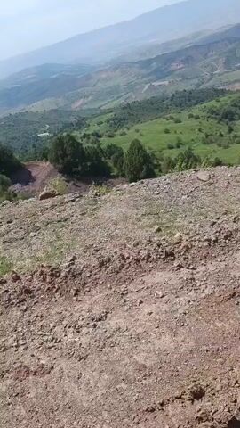 The video shows a drone flying over a mountainous, green landscape. In the foreground, there is a dirt path with rocks and sparse vegetation. The drone then moves forward, revealing a valley with more trees, green hills, and a winding dirt road or path. The sky is hazy and light blue, suggesting it is daytime and possibly a clear day with some atmospheric haze. The drone appears to be moving at a moderate pace, providing a sweeping view of the terrain. There are no people or animals visible in the video. The overall impression is of a natural, rugged environment.
