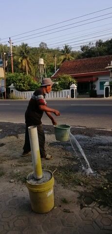 An elderly man is seen outdoors by the side of a road. He is wearing a dark shirt with red and white stripes around the collar and a cap. He is holding a green bucket in his right hand and pouring liquid into a white pipe that is sticking out of a yellow cylindrical object embedded in the ground. The pipe is secured in place by concrete. In the background, there is a road with a green truck passing by. Further in the background, there are houses, trees, and power lines. The sun is shining, indicating it is daytime.