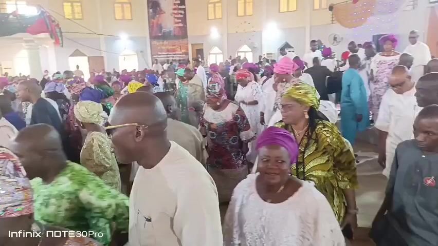 A large crowd of people are gathered in a large, bright hall. Many of the women are wearing colorful headwraps. Several people are wearing white. The hall is decorated with banners and streamers. People are moving around and smiling.