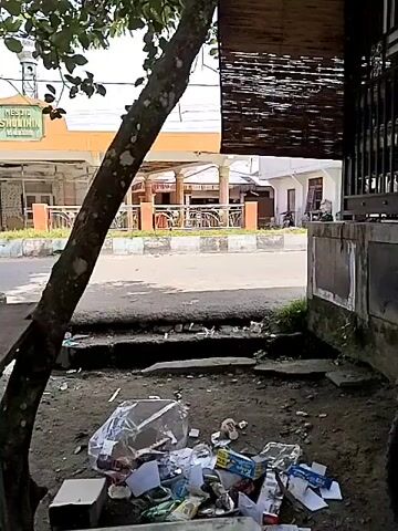 A woman in a green and white striped dress is sweeping a pile of trash with a broom. The trash includes plastic bags, wrappers, and other debris. She is standing on a dirt patch next to a tree. In the background, there is a street, a building with the words "Masjid Sholihin" written on it, and a bamboo blind. The scene appears to be outdoors in daylight.