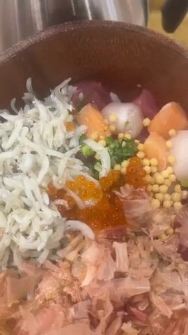 This is a close-up, top-down shot of a donburi bowl filled with various Japanese ingredients, likely a chirashi bowl, placed on a wooden tray. The bowl itself is a warm-toned, rustic wooden material. Dominating the bowl are layers of fresh ingredients: on the left, a generous pile of small, white, translucent fish, possibly shirasu (whitebait). In the center, a vibrant mound of bright orange ikura (salmon roe) glistens, surrounded by small yellow spheres, likely corn kernels or similar toppings. Interspersed are cubes of raw fish, including pink salmon and deep red tuna, along with white pieces that could be scallops or white fish. The bottom of the bowl is generously covered with a layer of thin, pinkish-brown flakes, which are katsuobushi (dried, fermented, smoked bonito flakes). A scattering of chopped green onions is visible throughout the toppings.

To the right of the donburi, a white ceramic dish contains a wooden spoon. In front of the donburi, two small white dishes are visible. The left one holds a dark liquid, likely soy sauce. The right one contains a green, paste-like condiment, possibly wasabi or a chopped herb mixture.

Behind the donburi, a brushed stainless steel thermal carafe with a black lid and handle stands tall. To the right of the carafe, a pair of white chopsticks rests on a paper wrapper. A faint hint of a white cup is visible in the background. The overall setting appears to be an indoor dining area, possibly a restaurant, given the presentation and the tableware. The lighting suggests it could be daytime. No people are visible in the frame, but the food is arranged invitingly, suggesting a meal about to be enjoyed. There is some Japanese text on the paper wrapper near the chopsticks, but it is too small and blurry to read.
