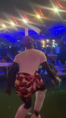 A man performs a traditional dance in a crowded nightclub. He is wearing a white t-shirt, a patterned skirt, white leg coverings, and a cap. He holds feather-like objects in his hands. The nightclub has colored lights, tables, chairs, and people in the background.