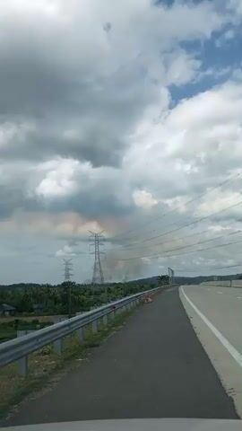 A video taken from inside a vehicle moving along a highway. The view is of the road ahead and to the right, with a clear sky and scattered clouds above. Several power lines stretch across the frame, supported by tall towers, one of which is visible in the distance on the left, with a plume of smoke rising near it. The highway has multiple lanes separated by white lines, and a metal guardrail runs along the left side of the road, separating it from a grassy area. There are no people or other vehicles immediately visible within the frame. The overall environment appears to be rural or semi-rural, with trees and vegetation visible beyond the guardrail.