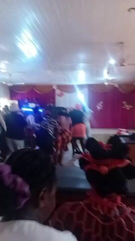 A group of people are dancing indoors in a room decorated with pink and white curtains. Some of the dancers are wearing brightly colored clothing, while others are wearing more subdued colors. A DJ booth is visible on a small stage at the end of the room. Bottles of water are sitting on a table in the foreground.