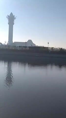 The video pans across a body of water, likely a lake or river, with a clear blue sky overhead. In the distance, various structures are visible. On the left, a tall, slender tower with a decorative top stands out. Further back, a building with a distinctive roof shape is present. Along the shoreline, there is a walkway with lampposts and trees. In the latter part of the video, more buildings and a line of people are visible along the waterfront. The water itself is calm, with reflections of the sky and the distant structures. The overall impression is of a park or recreational area on a clear day.