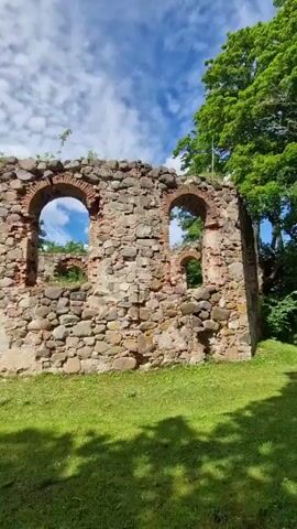 The video shows the exterior of ancient stone ruins of a castle on a sunny day. The ruins are made of large stones and bricks, with arched windows and doorways. Green grass covers the ground around the ruins, and lush green trees are visible in the background. The sky is blue with white clouds. The camera pans across the ruins, showing different angles and parts of the structure, including a tall, intact tower. There are no people or animals visible in the video. The scene is peaceful and historic.