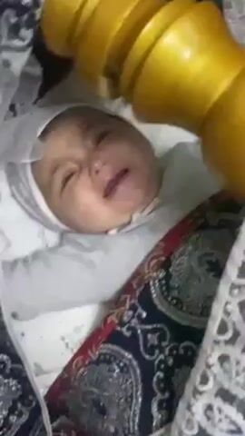 A close-up, slightly angled, medium shot shows a baby lying in a crib. The baby, wearing a white bonnet and white clothing, is crying with a distressed expression. The crib is covered with a patterned fabric in colors of blue, red, white, and gold. Above the baby, slightly out of focus, is a yellow object. The camera is held at an angle, offering a direct view of the baby's face.