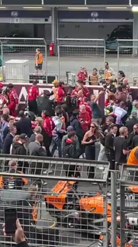 The video shows a large crowd of people milling around in front of a garage at what appears to be a racetrack, with several team members in red Ferrari racing uniforms visible. The crowd is a mix of individuals in formal attire, team uniforms, and casual clothing, all standing behind a safety fence. A bright orange race car is partially visible behind the fence, and DHL logos can be seen at the top of the garage doors. A man in a dark jacket throws his arms up in the air as if in celebration. The sound of a race car engine can be heard in the background.