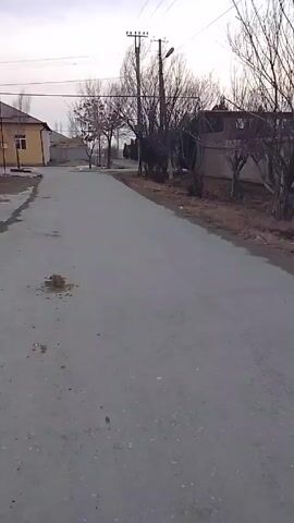 The video shows a sparsely populated residential street in what appears to be a rural or suburban area. The street is asphalt and has some minor damage. Along the side of the street are houses, some of which are under construction. Leafless trees and bushes border the road. Utility poles with overhead wires run along the side of the street. The sky is overcast, suggesting a dull or late-day lighting. There are no people or vehicles immediately visible in the frame.
