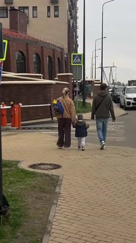 A family of three is walking across a crosswalk. They are walking away from the camera. The crosswalk is made of tan brick pavers. To the left is a brick building. To the right is a street with parked cars. The sky is overcast. The crosswalk sign is visible. The mother is wearing a patterned sweater and brown pants. She is holding the child's hand. The child is wearing a blue vest and a grey hat. The father is wearing a green hoodie and jeans. He is carrying a water bottle. They are all walking at a moderate pace. The camera is stationary and focuses on them as they cross.