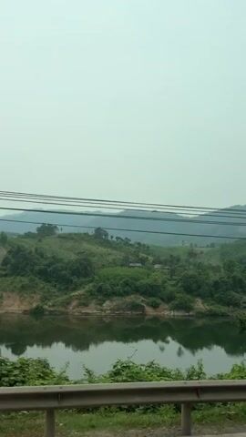 The video shows a view of a landscape from a moving vehicle, likely a car. The scene includes a body of water, possibly a river or lake, surrounded by lush green vegetation and trees. The water reflects the sky, which appears overcast or hazy. In the distance, there are rolling hills or low mountains. Power lines run across the top of the frame. A metal guardrail is visible in the foreground, suggesting the vehicle is on a road or highway. The camera pans slightly, focusing on different parts of the landscape as the vehicle moves.