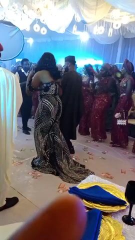 A group of people are dancing at a formal event. The camera is focused on a woman in a sequined, strapless, floor-length gown, and a man in a dark suit and cap. The woman is dancing with her back to the camera, and the man is facing her. Other guests, mostly women in red traditional attire, are visible in the background. The room is decorated with white fabric draping from the ceiling and lit by hanging lights. There are gold and blue plates on a table in the foreground.