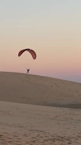 A paraglider is seen soaring over sand dunes against a gradient of pink and orange sky during what appears to be sunset. The paraglider, small in the frame, is suspended in mid-air, attached to a colorful parachute-like wing. The vast expanse of sand dunes stretches across the lower portion of the video, marked with footprints and tire tracks. In the background, distant structures and vegetation are visible near the coastline, with the ocean meeting the horizon. The overall scene conveys a sense of freedom and adventure in a natural, arid environment.
