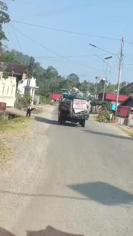 The video shows a view from inside a vehicle traveling on a paved road. A truck loaded with what appears to be construction materials, including metal scaffolding and wood, is in front of the vehicle. The truck has a white sign on its back with writing in what appears to be Indonesian. To the left of the road, there are buildings and vegetation. A person is walking on the side of the road. Further down the road, a motorcycle is visible. The road is surrounded by trees and hills in the background. The sky is clear and the sun is shining.