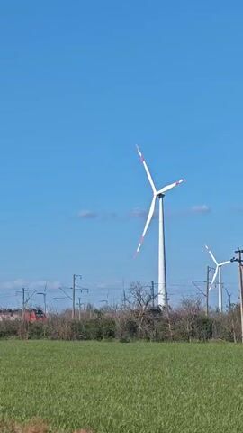 The video shows a wide shot of a grassy field under a clear blue sky. In the mid-ground, two wind turbines are visible. The larger turbine on the left is closer to the camera and its blades are slowly rotating. To the right, a smaller turbine is also visible with its blades rotating. Behind the turbines, a train with red and black carriages is passing by on railway tracks, obscured by a line of trees and bushes. There are also power lines and poles in the background. The overall impression is a rural landscape with industrial elements.