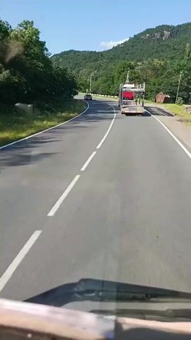 A vehicle is traveling on a road, passing a red car on its left. The road is paved and has white lines marking the lanes. In front of the vehicle, a red car carrier truck is also traveling on the road. The truck is empty. The landscape is green and mountainous, with trees lining the sides of the road. The sky is blue and clear. The scene appears to be a sunny day.