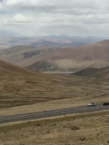 The video shows a mountainous landscape, presumably the Himalayas. The foreground is a slope covered in dry, brown grass. In the middle ground, a winding road snakes through the valley, with a white car driving along it. In the background, there are layers of mountains in various shades of brown and grey under a cloudy sky. The weather seems overcast, but visibility is clear enough to distinguish the details of the terrain and the vehicles on the road. The camera appears to be stationary.