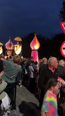 A crowd of people are gathered at dusk to watch a parade float. The float is shaped like a bulbous pink and yellow tower topped with a gold finial and illuminated from within. To the left of this float, a similar float is visible, featuring a person in a golden outfit sitting in a boat-like structure under a golden umbrella. The crowd is diverse in age and gender, with many holding up phones to capture the event. Traffic signs, including a yield sign and a circular sign with an arrow, are visible on the right. Overhead, paper lanterns in the shapes of stars and a crescent moon hang from wires. The background consists of trees and a dark sky, indicating the time of day is evening.