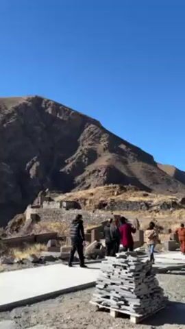 A landscape shot shows a group of people walking along a path toward a stone structure undergoing construction. Scaffolding surrounds the building. In the foreground, a stack of stone tiles is visible. A rugged mountain range forms the backdrop under a clear blue sky.