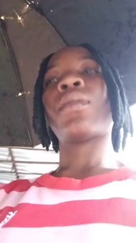 A close-up video shows a young Black person with braided hair wearing a red and white striped shirt. The person is holding an umbrella, as evidenced by the dark fabric visible above their head. The person's face is the primary focus, and they appear to be looking in different directions. The lighting suggests an outdoor environment, possibly during daytime.