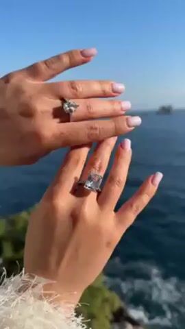 The video shows two hands, each with a large engagement ring, held up against a backdrop of a blue sea and sky. The rings are different styles; one appears to be a pear-shaped diamond, and the other is a large, rectangular-cut stone. The fingers have manicured nails with a light pink polish. The person wearing the rings is standing outdoors, possibly on a cliff or elevated point, as there is vegetation visible at the bottom of the frame. The focus is on the rings and the hands displaying them.