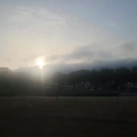 The video shows a sunrise over a landscape of hills and trees. The sun is partially obscured by clouds, casting a bright glow. In the foreground, there is a dark area that appears to be grass or dirt. There are some indistinct shapes that might be vehicles on a road in the middle ground. The overall impression is one of a peaceful morning scene.